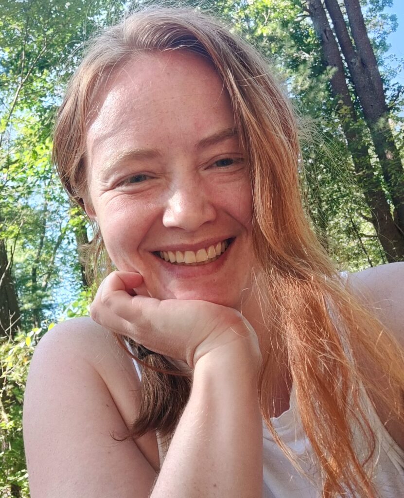 Writer H. Halliday Nelson - poet, author, blogger, and content creator. She is sitting outside with trees behind her and most of her long red hair pulled back. She is resting her chin on her hand and smiling at the camera.